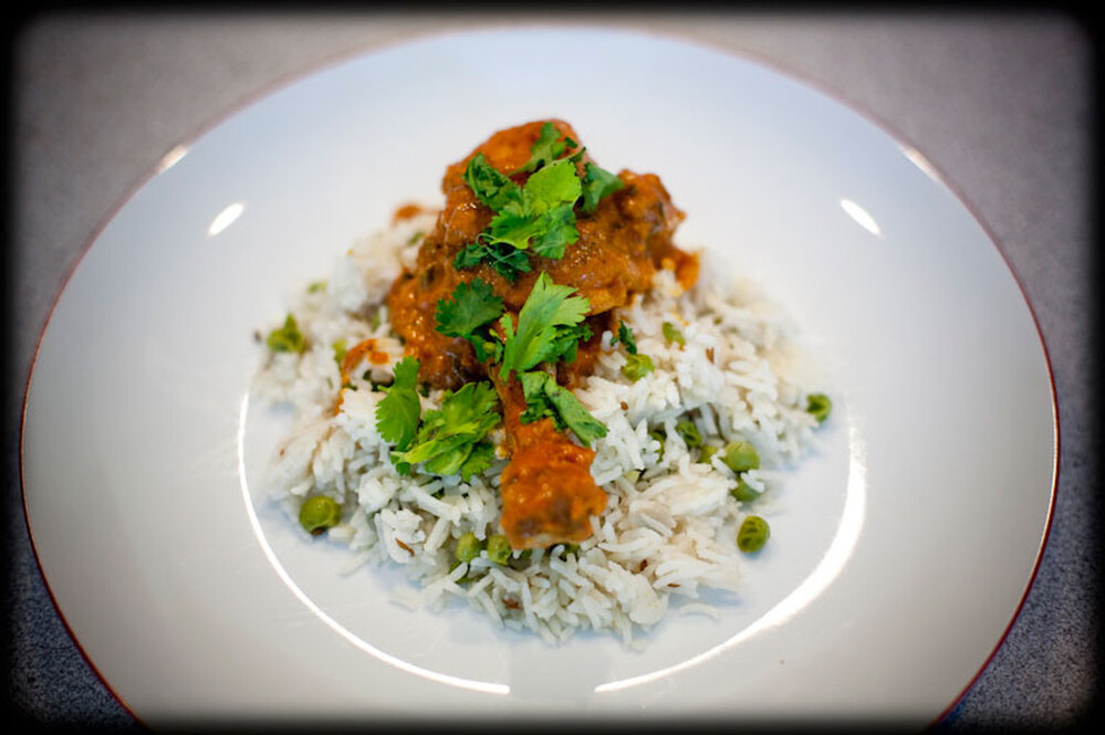 A piece of butter chicken rests on a bed of rice and peas, with a garnish of greens