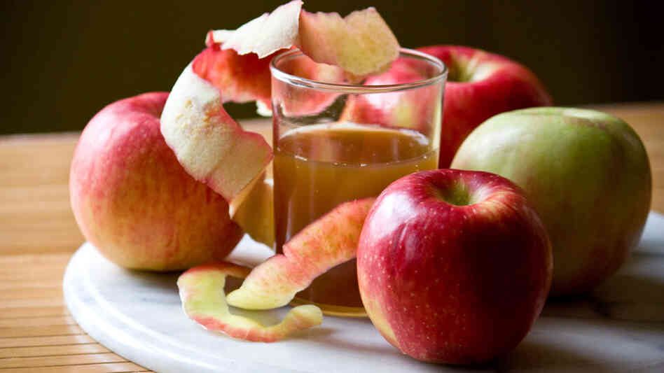 A glass of apple cider is nestled among apples of varying hues on a marble charger
