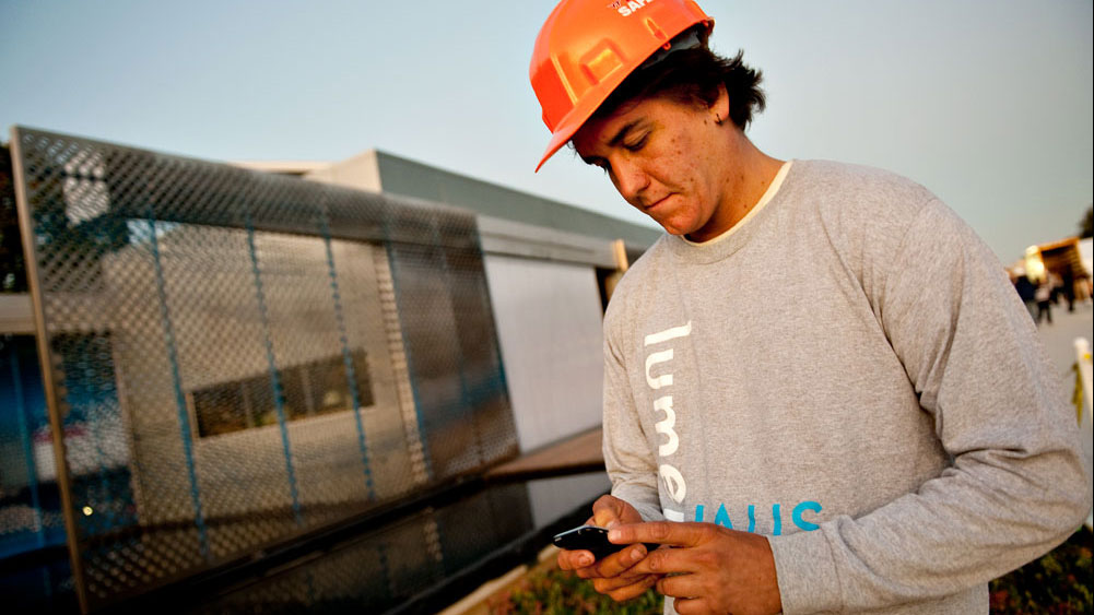 An architecture student demonstrates the iPhone, with the solar home in the background.