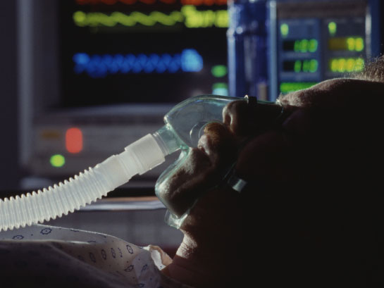 A man breathing with the help of a ventilator lies in a hospital bed.