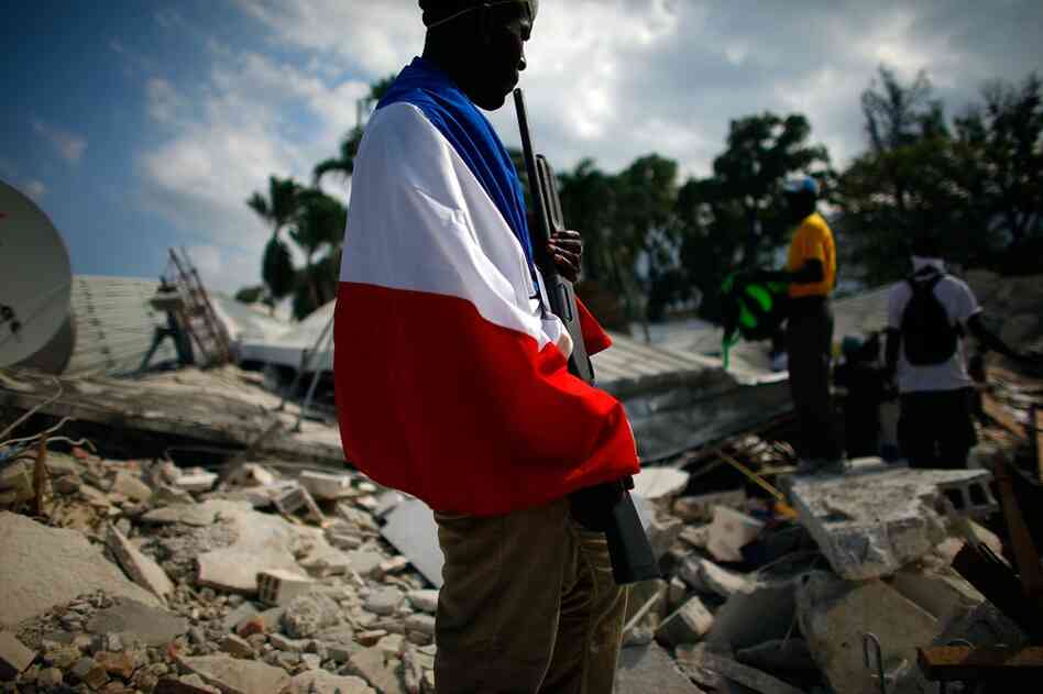 A Haitian man, draped in a French flag, guards a government building with a shotgun near the Port-au-Prince waterfront.
