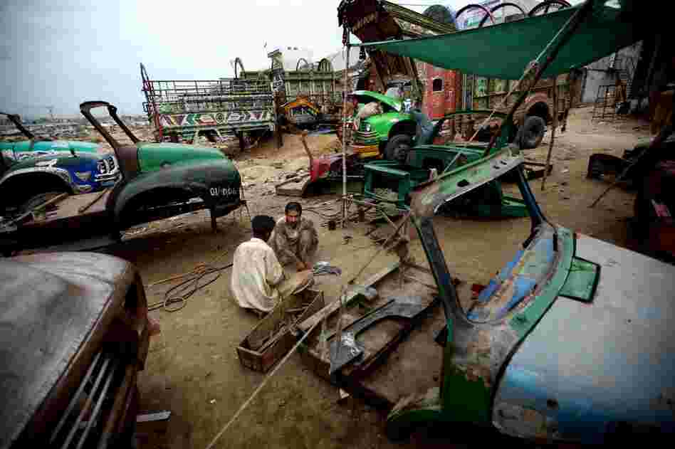 At the makeshift home of a large gathering of mechanics, welders, craftsmen, painters and metalworkers — all working on a fleet of distinctive Pakistani transport trucks — workers shape metal with hammers amid old truck cabs. In the background, mechanics work on a truck engine.