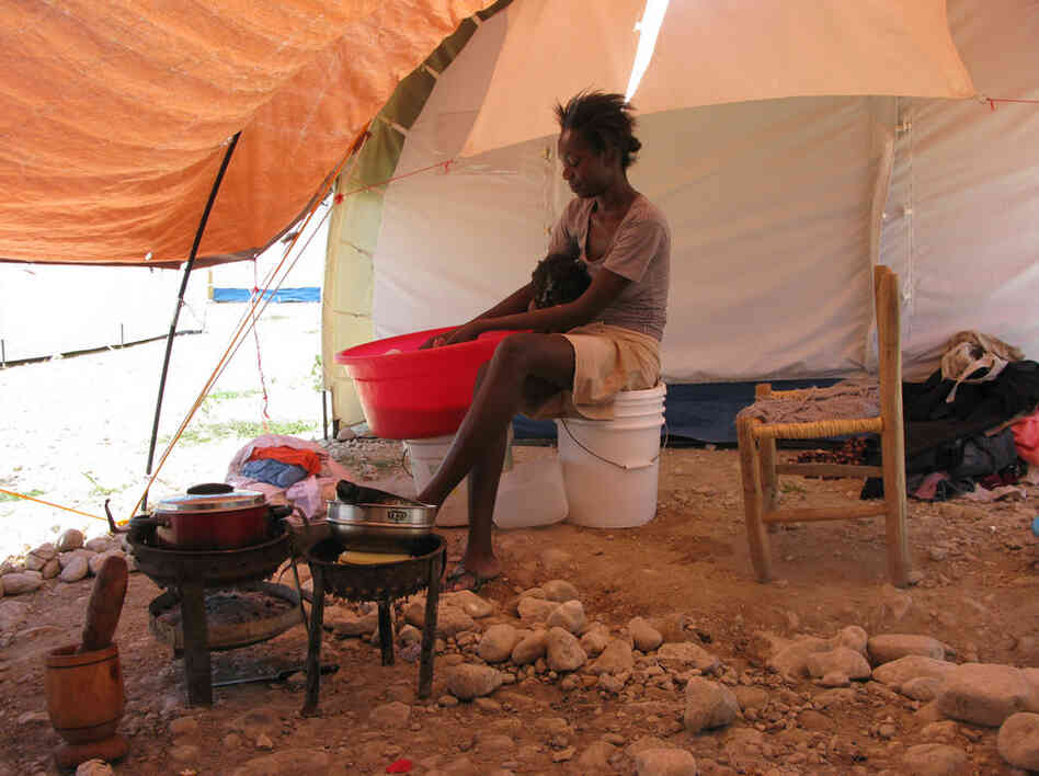 A Haitian woman cooks beans and washes clothes outside her tent. A tarp protects her and her daughter from the scorching sunlight. 