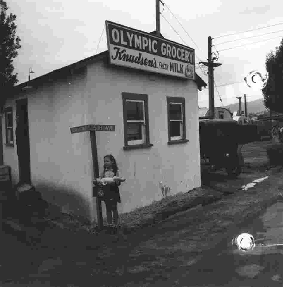 A young girl, seen with her baby doll, stands outside of Olympic Grocery in Santa Monica. Some residential trailers can be seen in the background on the right.