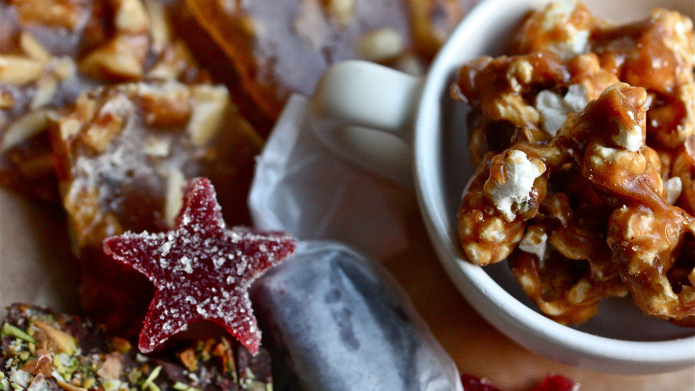 An assortment of homemade candies, including a sugared red jelly star, toffee, nut brittle, a caramel wrapped in parchment paper and a mug full of caramel corn.