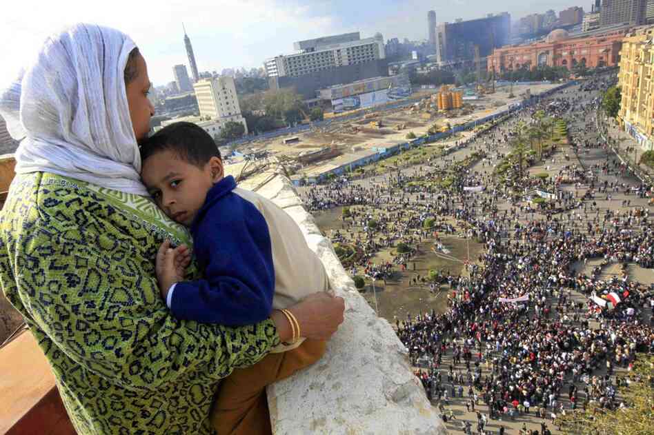 A woman hugs her child as she watches some thousands of protesters gather at Tahrir square. In the distance, the ruling National Democratic party building smolders behind the red-colored Egypt museum. 