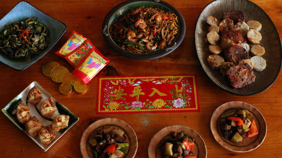 A wood table set with Chinese New Year decorations and a Trinidadian-Chinese feast, with wontons and stir-fried meats and noodles
