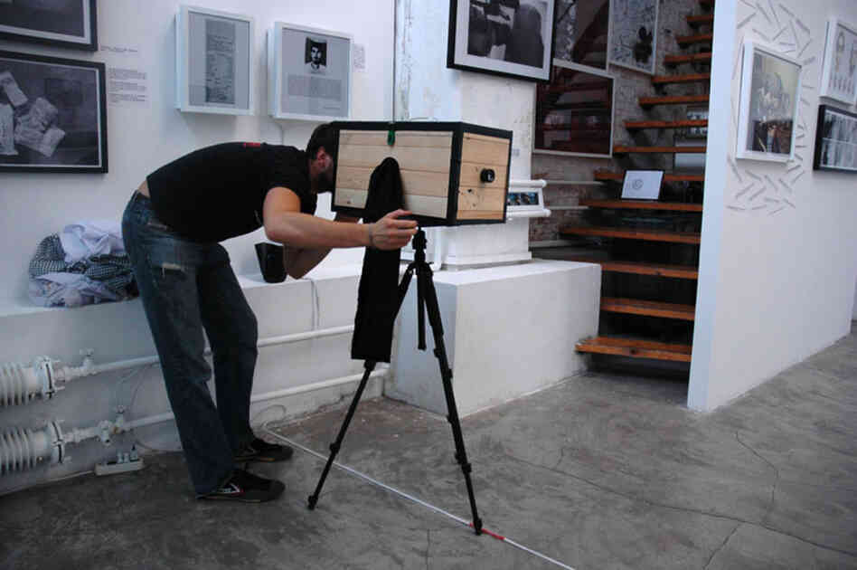 Birk peers through the open back of his box camera. To focus the camera, he moves a translucent glass plate toward or away from the lens until the image projected on the glass is sharp. This is where he will mount the photo-sensitive paper once the light is sealed out.