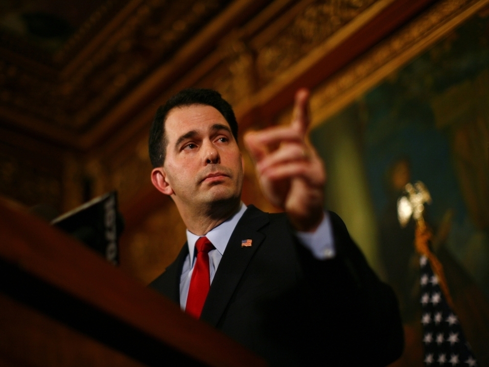 Republican Gov. Scott Walker speaks at a  news conference inside the Wisconsin State Capitol in Madison on Feb. 21 while people protest the legislation proposed by Walker to restrict collective bargaining.
