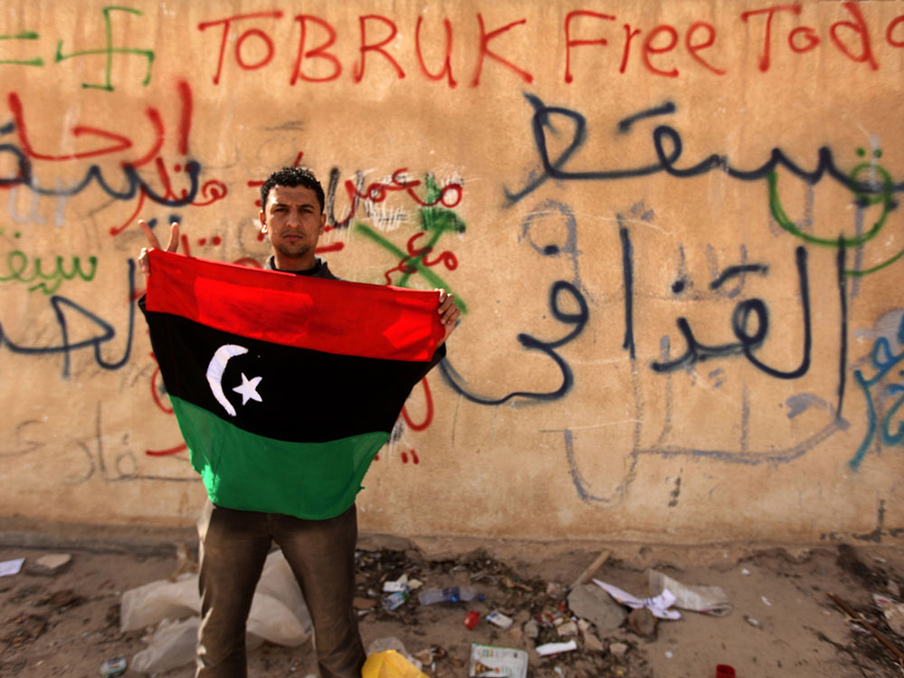 A protester holds an old national flag in front of a wall covered with graffiti against Libyan leader Moammar Gadhafi in the eastern city of Tobruk on Thursday. A protester holds an old national flag in front of a wall covered with graffiti against Libyan leader Moammar Gadhafi in the eastern city of Tobruk on Thursday.