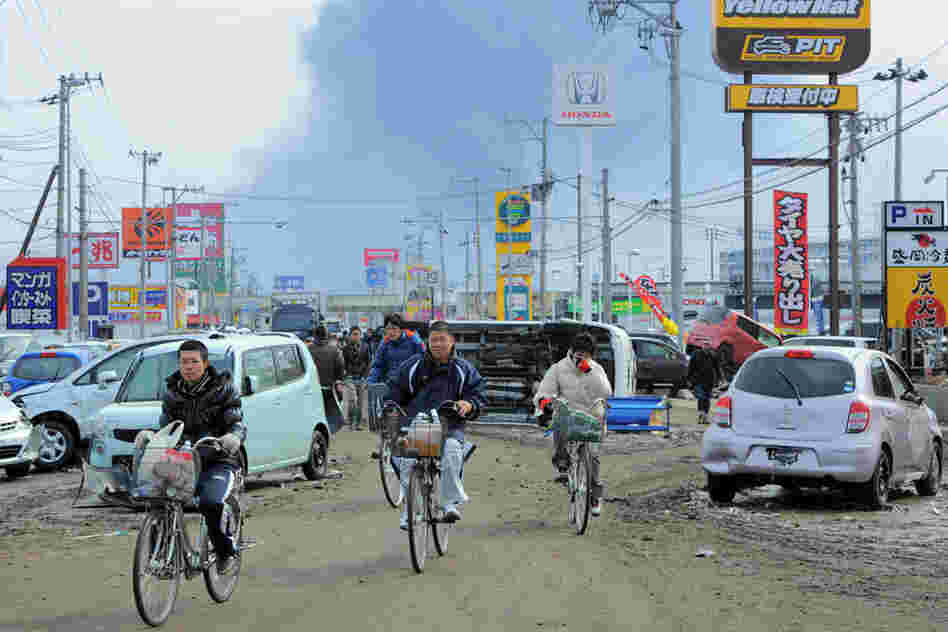 Local residents cycle past damaged cars littering a street in Tagajo, Miyagi prefecture, as fire smoke rises in the distance. 