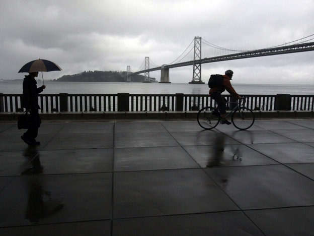 A pedestrian and cyclist are seen along the Embarcadero with the San Francisco Oakland Bay Bridge in the background. San Francisco dismantled a freeway in this location in the early 1990s.