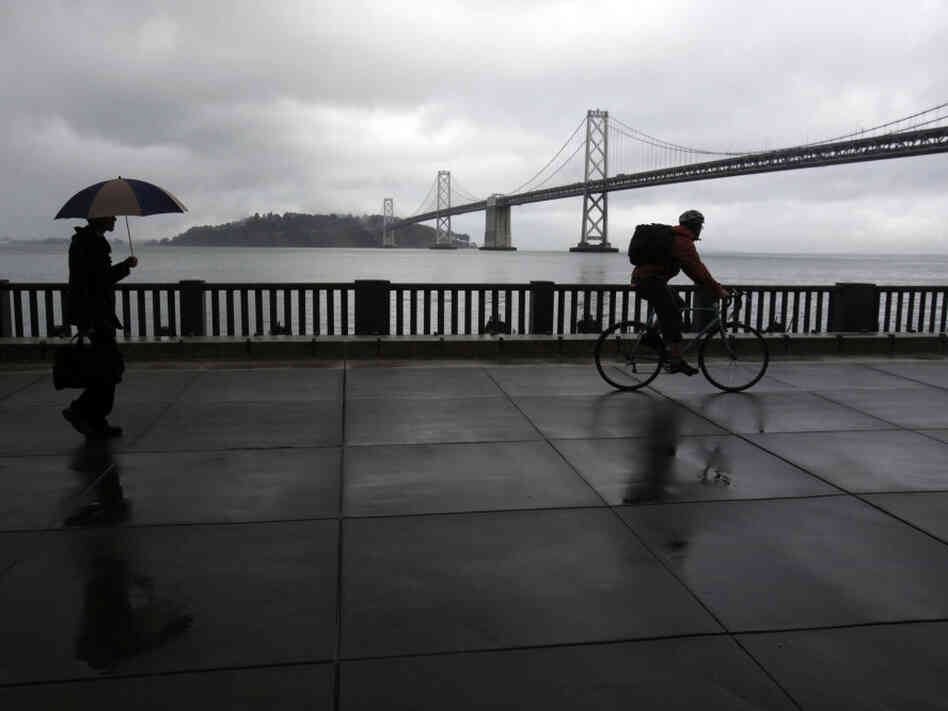 A pedestrian and cyclist are seen along the Embarcadero with the San Francisco Oakland Bay Bridge in the background. San Francisco dismantled a freeway in this location in the early 1990s.