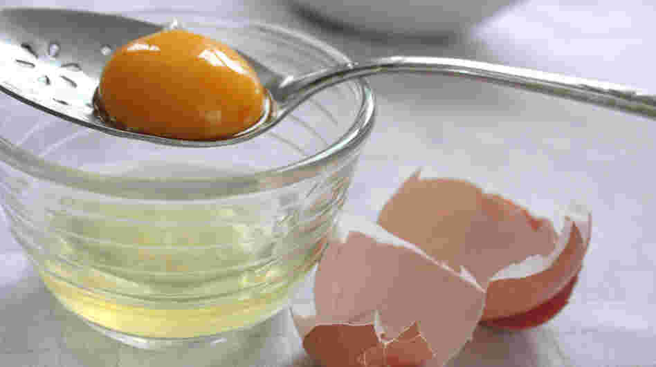 A yolk rests in a slotted spoon over a bowl containing the separated egg white, with the egg shell cracked alongside