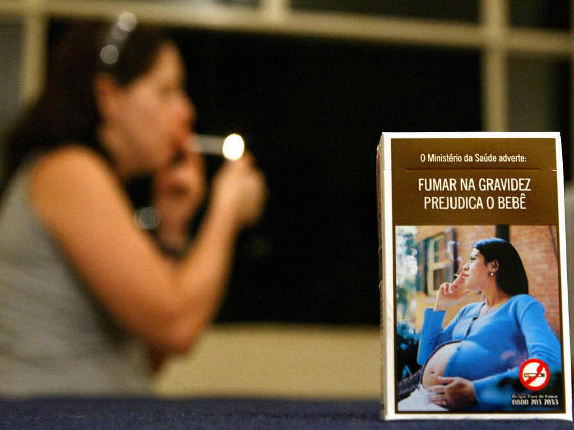 A Brazilian cigarette pack shows a pregnant smoker with the caption "The Ministry of Health warns: smoking during pregnancy is detrimental to child's health," while a woman lights a cigarette in the background.