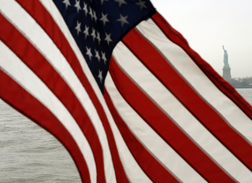 The American Flag blows in the wind off the back of the Ellis Island/Liberty Island Ferry with the Statue of Liberty in the background.