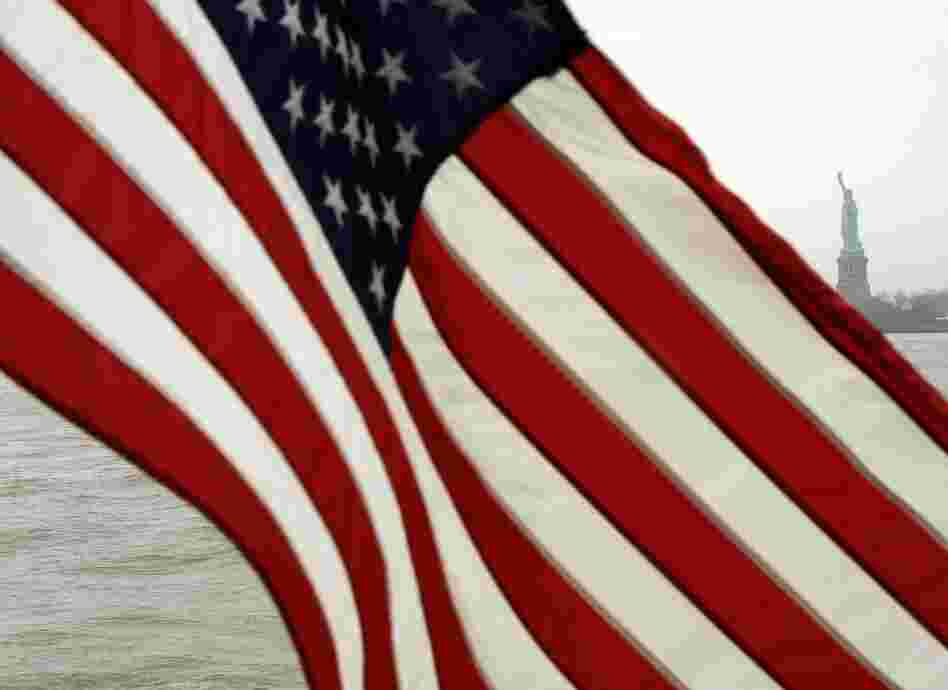 The American Flag blows in the wind off the back of the Ellis Island/Liberty Island Ferry with the Statue of Liberty in the background.