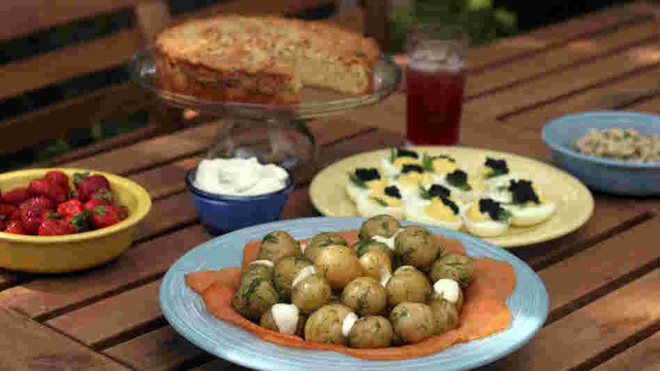 A misdsommar feast spread on a picnic table includes cake and strawberries, new potatoes and smoked salmon, shrimp salad and a special cocktail.