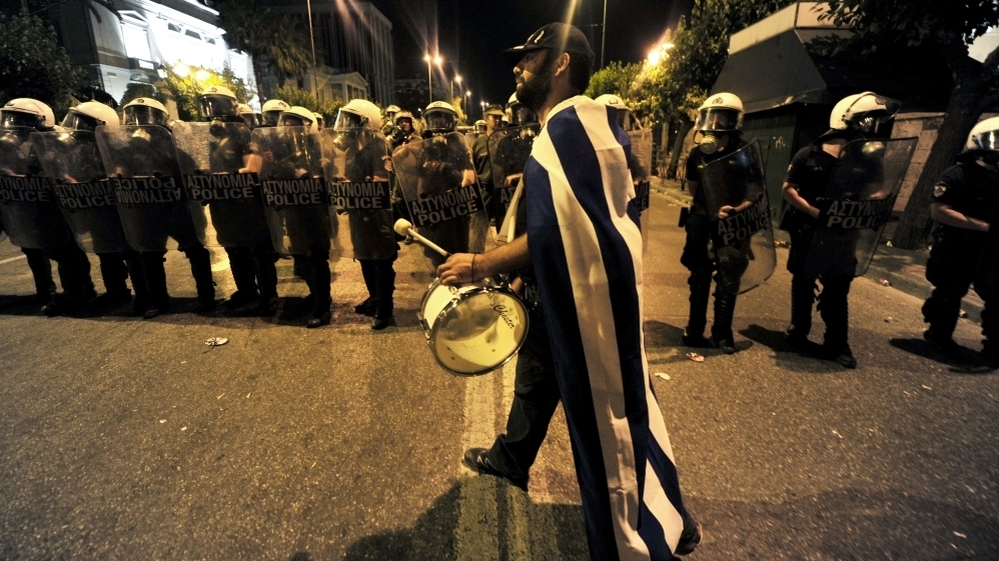 A demonstrator plays drum in front of riot police outside the Greek Parliament in Athens. The protesters aren't the usual leftists and trade unionists with red banners. They're mostly middle-class Greeks waving the white and blue national flag.