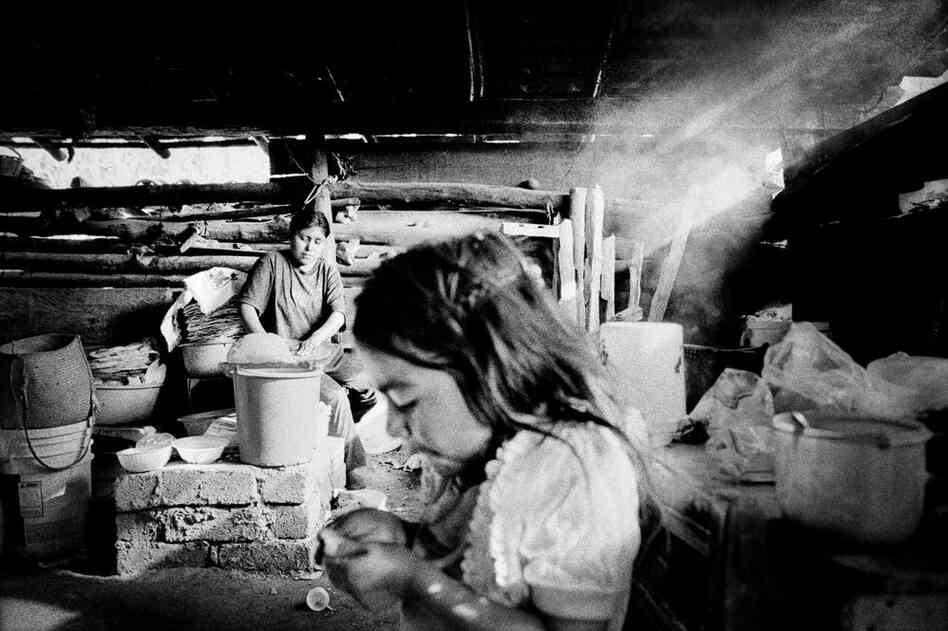 A woman makes tortillas on an indoor fire while her daughter plays nearby.