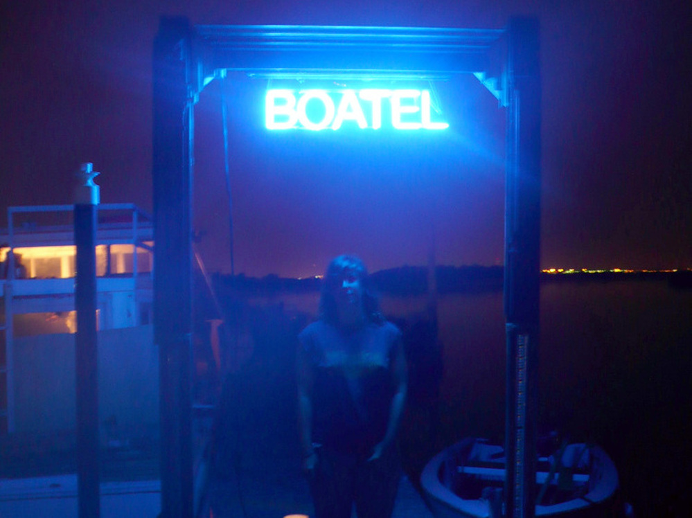 Constance Hockaday, the creator of the Boatel, stands under the sign announcing her art project/hotel at Marina 59 on Jamaica Bay in Queens, New York.