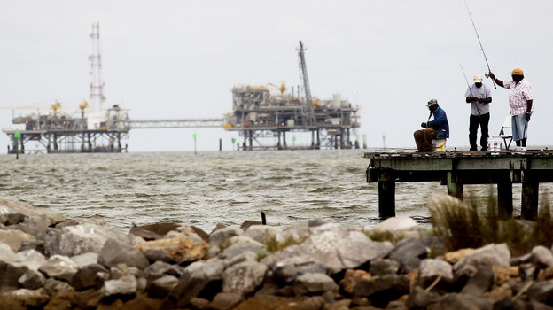 Men fish off a pier at a jetty in Dauphin Island, Ala., with oil rigs in the background. The U.S. government is changing how it regulates drilling platforms.