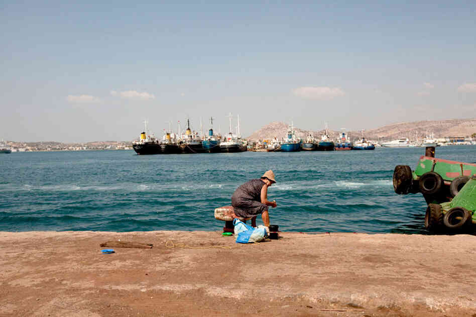A woman fishes off the Perama coast. The ships behind her, waiting for repair, have been abandoned.
