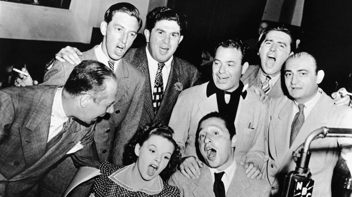 Bert Lahr (far right), Ray Bolger (back row, right), Judy Garland (sitting, right), composer Harold Arlen (sitting left), and various MGM and music publishing executives sing songs from the 1939 film musical 'The Wizard of Oz' around a microphone in the NBC radio studio, circa 1939. 