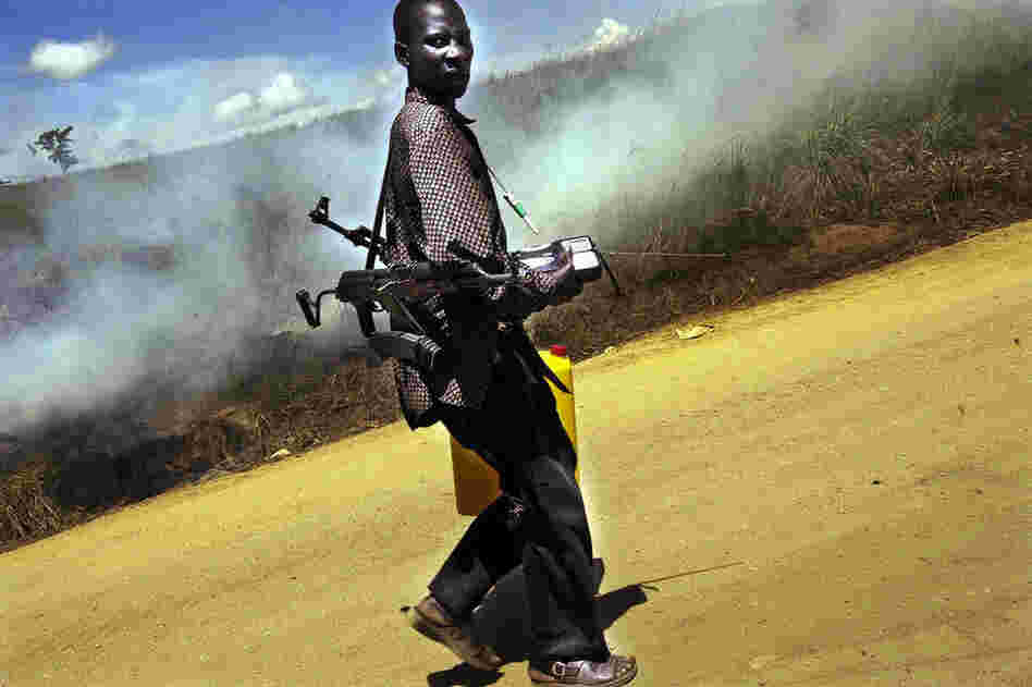 A Congolese fighter carries two AK-47s past burning bushes following strife in the Ituri region of the Democratic Republic of Congo in 2003. 