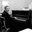 Jerry Lee Lewis smiles over his shoulder while playing the piano in a dressing room.