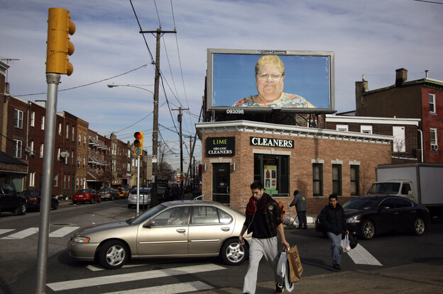 Pedestrians in Philadelphia cross a street in view of a billboard with a photo by Zoe Strauss. Only a dozen years after first picking up a camera to chronicle her beloved hometown's overlooked people and places, Strauss was honored with a solo show at the Philadelphia Museum of Art.