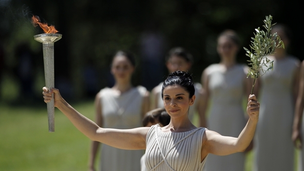A priestess holds an olive branch and the lit Olympic torch.