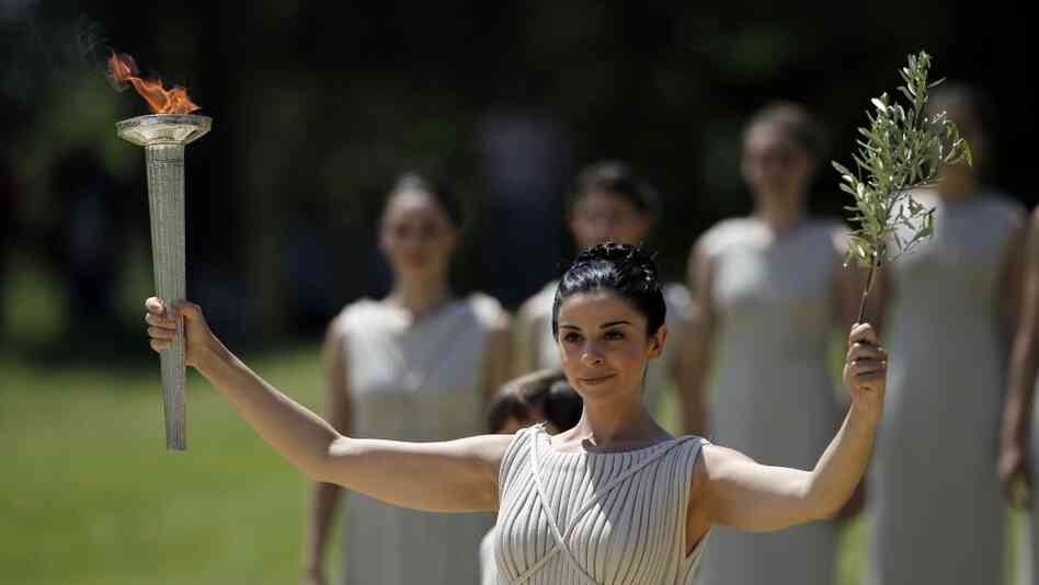 A priestess holds an olive branch and the lit Olympic torch.