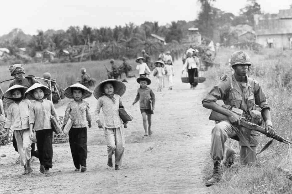 An American soldier guards the road as Vietnamese women and schoolchildren return home, December 1965.