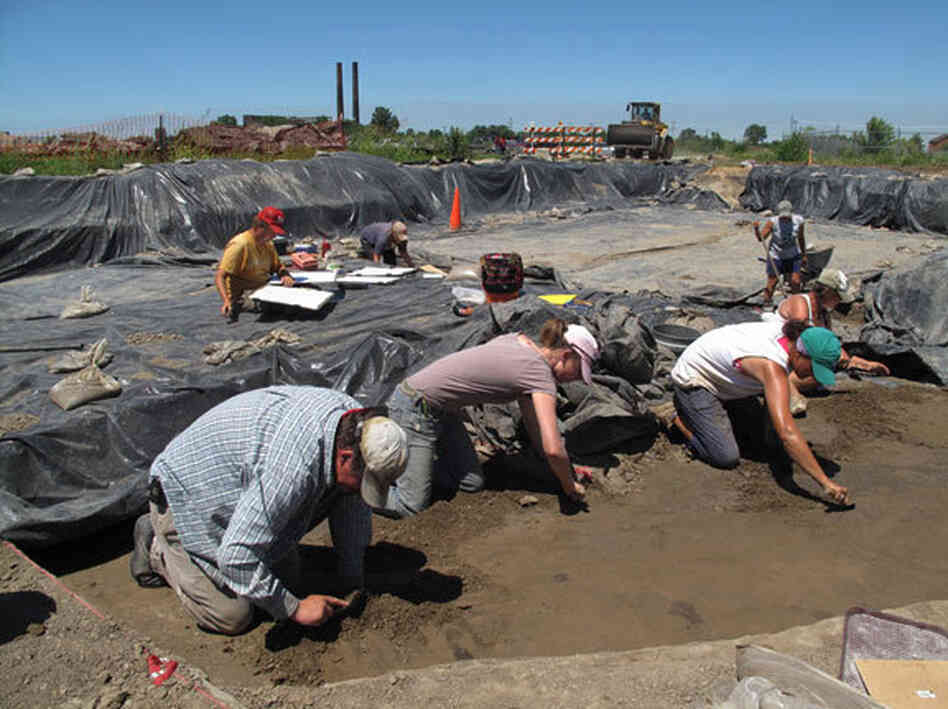 Illinois State Archaeological Survey crew members scrape the soil in search of Cahokian artifacts. The crumbling smokestacks of a 19th century meat packing plant are visible in the background, along with a bulldozer working on the current interstate freeway construction.