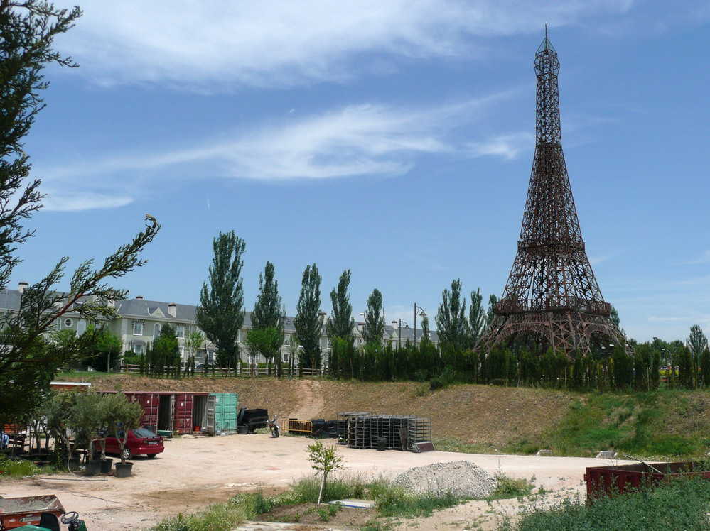 A rusting model of the Eiffel Tower stands at one end of the theme park, surrounded by construction debris and empty housing outside the park.