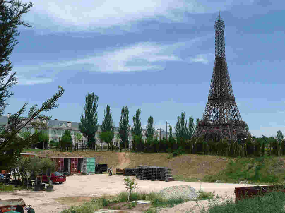 A rusting model of the Eiffel Tower stands at one end of the theme park, surrounded by construction debris and empty housing outside the park.