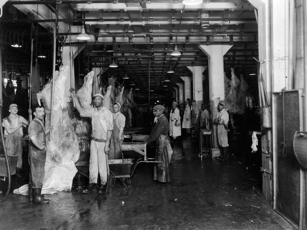 Men at a slaughterhouse stand near hanging beef carcasses, late 1940s.
