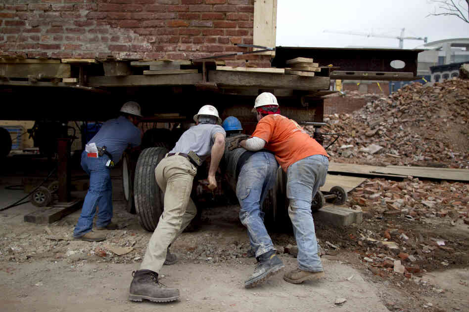 Workers position platforms with tractor-trailer-size tires under a building.