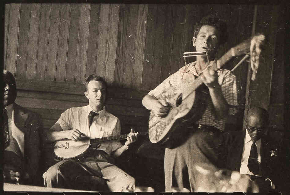 Guthrie plays his signature guitar with "This machine kills fascists" scrawled across the front. Pete Seeger accompanies on banjo; music journalist Dan Burley sits at left.