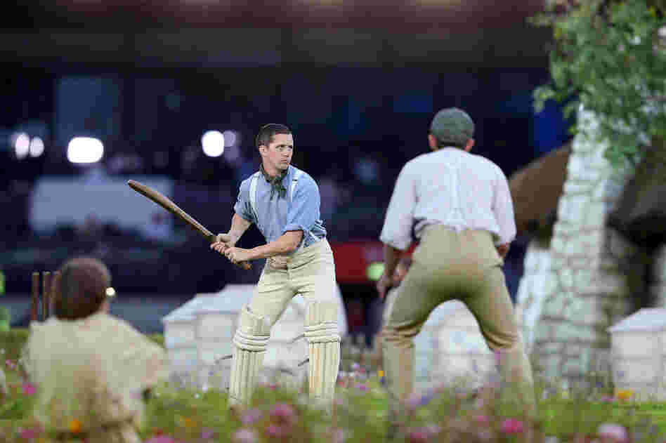 Performers depict a view of the English countryside by playing a game of cricket.