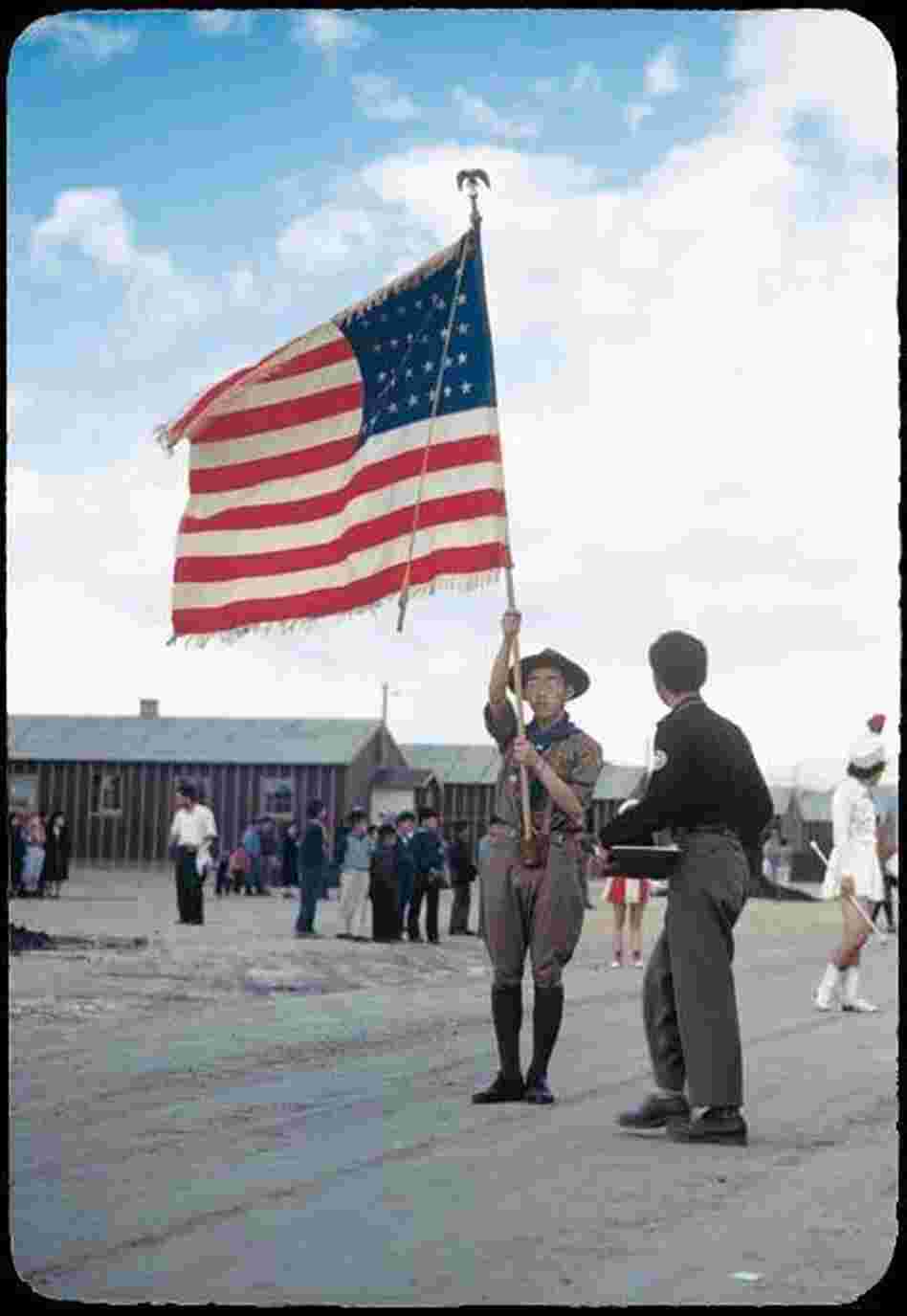 A Boy Scout carrying an American flag leads a parade through the Heart Mountain camp.
