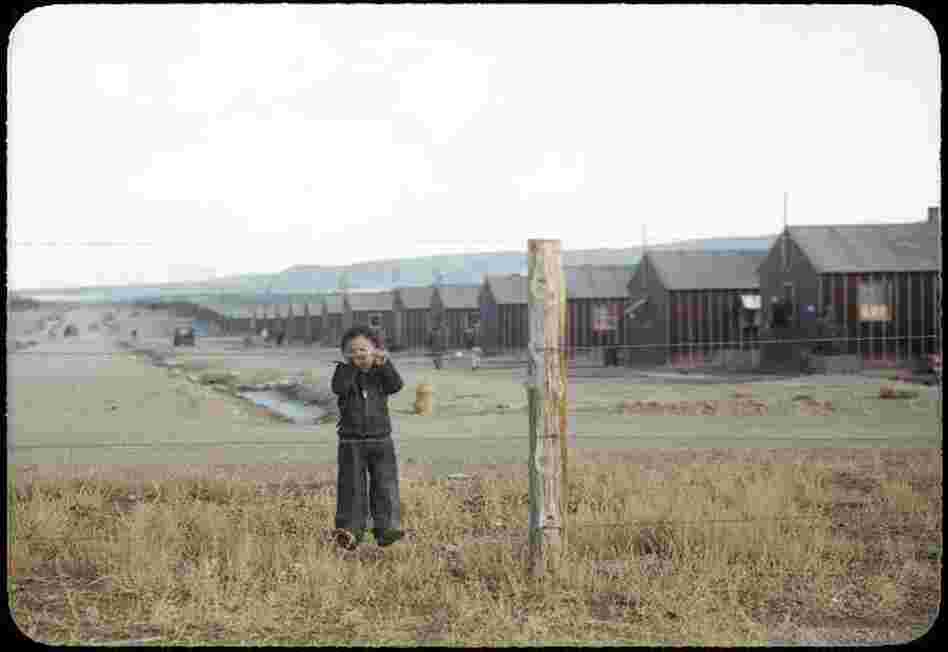 Billy Manbo clings to a barbed-wire fence enclosing a row of tar paper barracks.