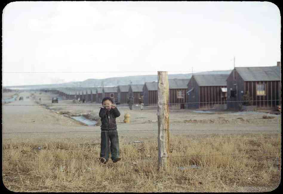 Billy Manbo clings to a barbed-wire fence enclosing a row of tar paper barracks.