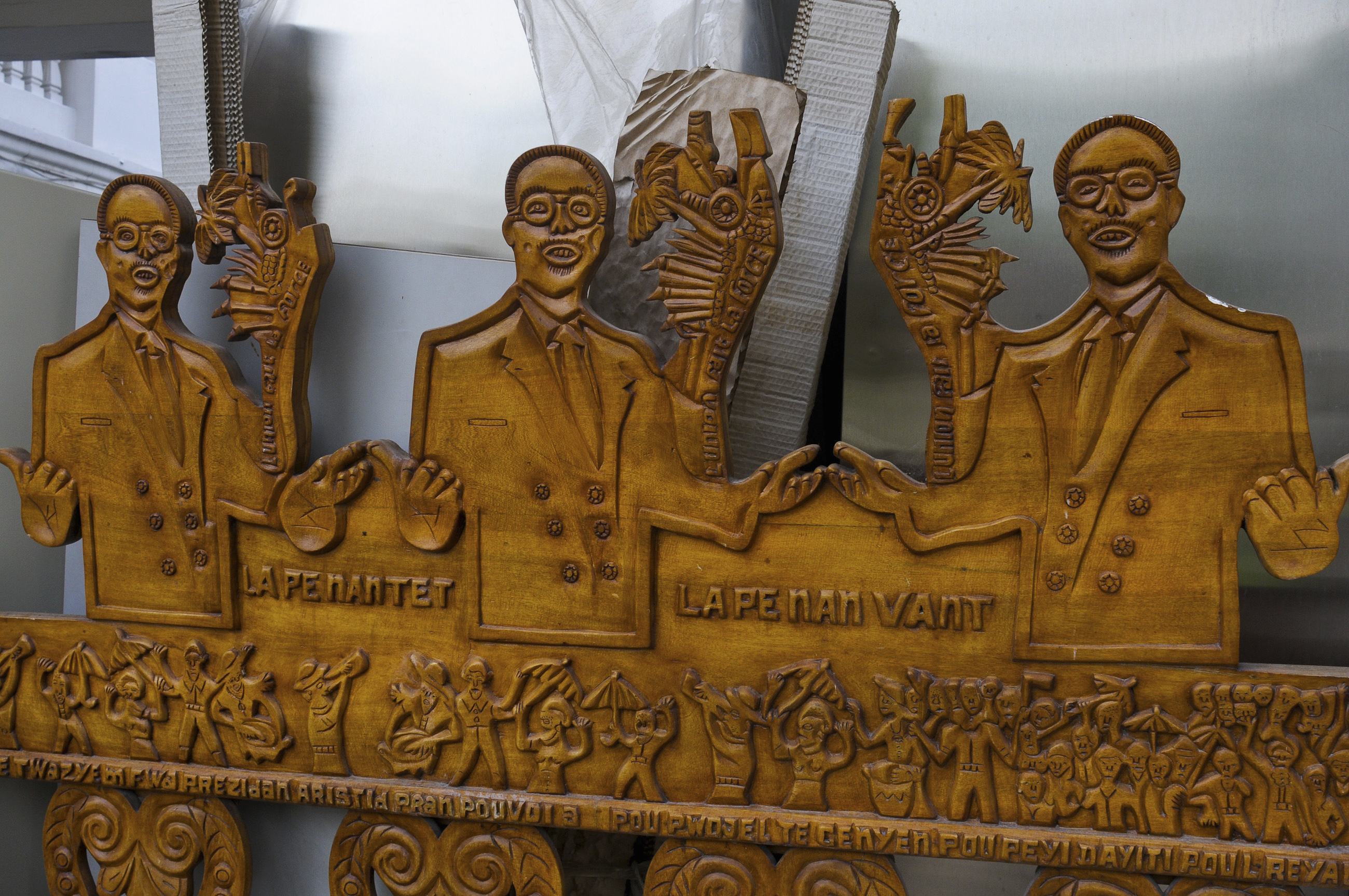 A wooden sculpture depicting former Haitian President Jean-Bertrand Aristide rests against a wall of the National Palace. The words on the sculpture that read 