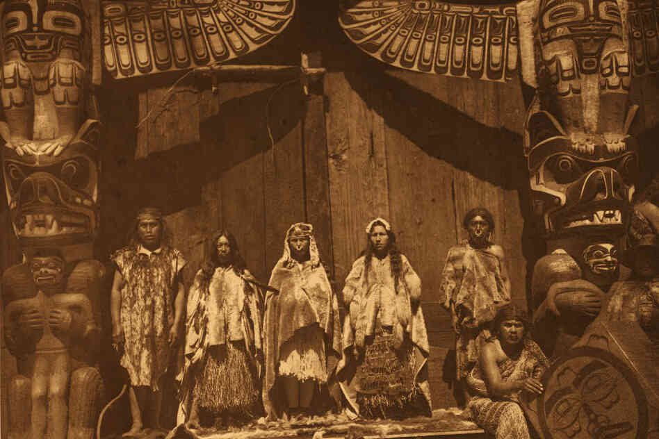 The original caption reads: "The [Kwakiutl] bride stands in the middle between two dancers hired for the occasion. Her father is at the left, and the bridegroom's father at the right behind a man who presides over the box-drum."