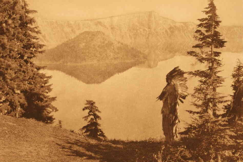 For a photo simply titled "Chief — Klamath," the caption reads: "The subject of this plate, in deerskin suit and feathered war-bonnet of the Plains culture, is shown against a background of Crater lake and its precipitous rim towering a thousand feet above the water."