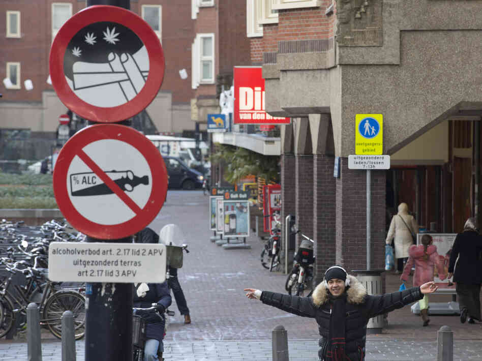 A young man with a joint in his left hand jokes with the photographer as he passes a sign prohibiting the use of marijuana and alcohol, in a designated area in Amsterdam on Wednesday. The city's mayor says students would be formally banned from smoking marijuana at school as of Jan. 1, 2013.