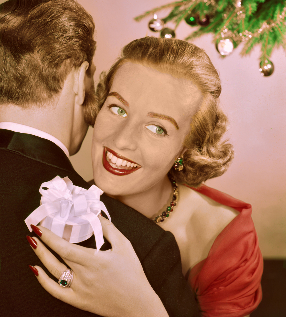 A lady hugs a man whilst she holds onto a Christmas gift wrapped in a bow on December 25, 1955.