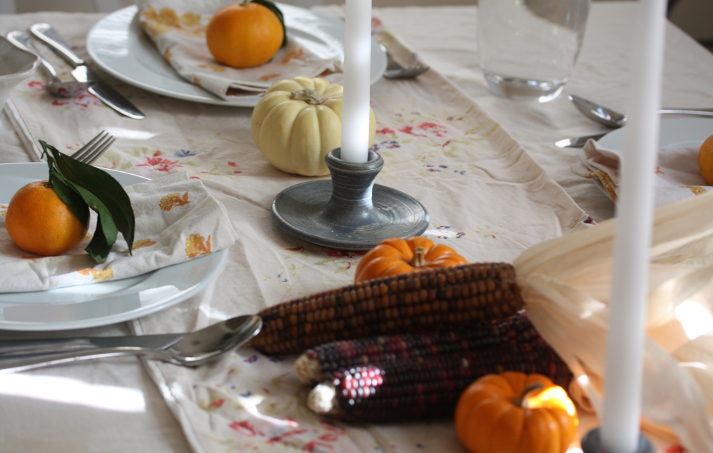 A dining table set for the holidays with linens, candlesticks, clementines and gourds.